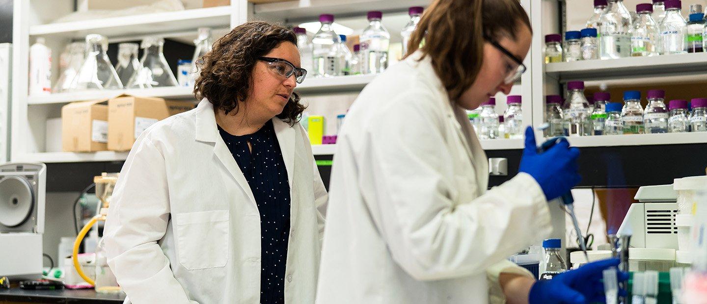 Two women in a lab with chemicals in glass jars.